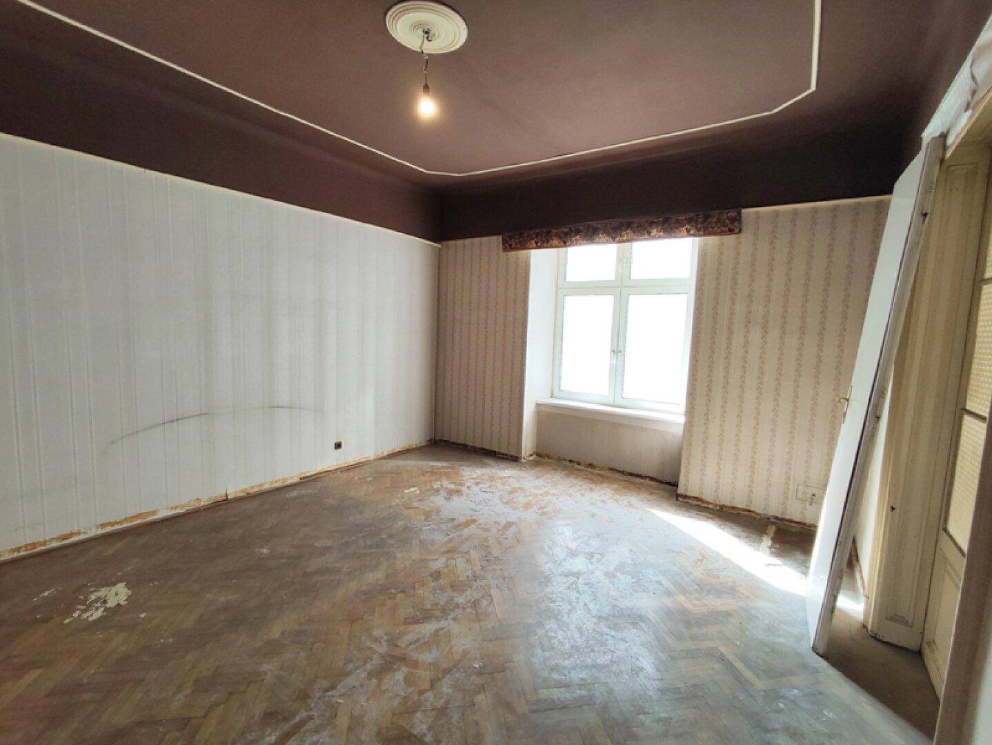 Innenraum mit abgenutztem Fischgrätparkett, brauner Decke und einem großen Fenster.