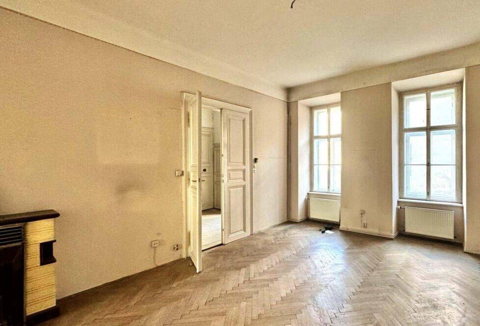 Helles Wohnzimmer mit Fischgrätparkett, zwei Fenstern und einem Ofen für gemütliche Abende.