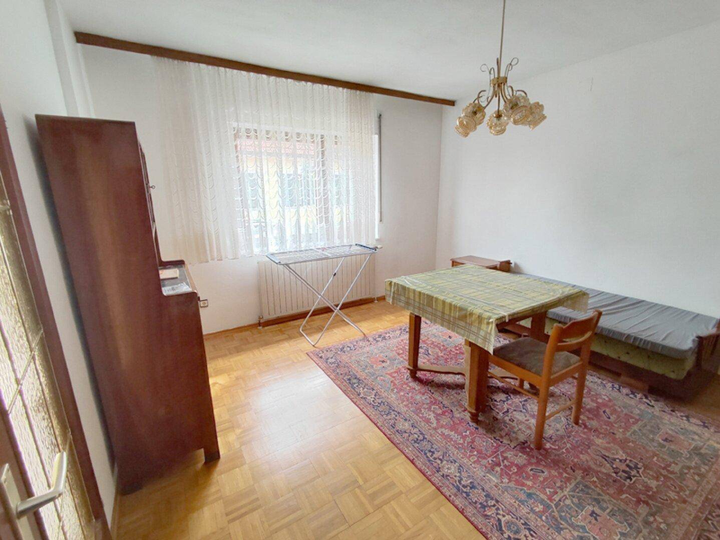 Einfaches Zimmer mit Parkettboden, Tisch und Fenster mit Vorhängen.