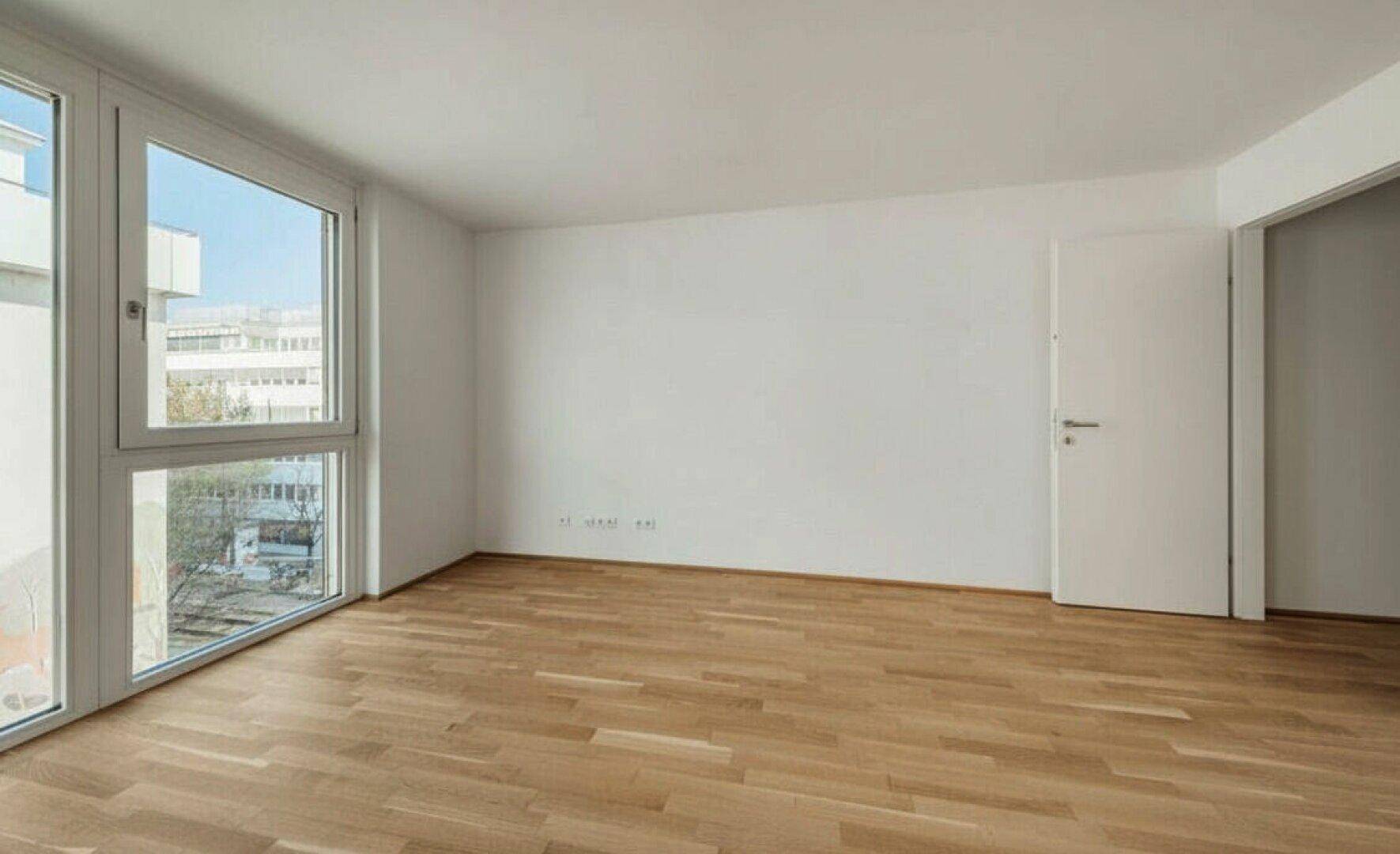 Helles, leeres Zimmer mit Holzboden und großen Fenstern, die viel Tageslicht hereinlassen.