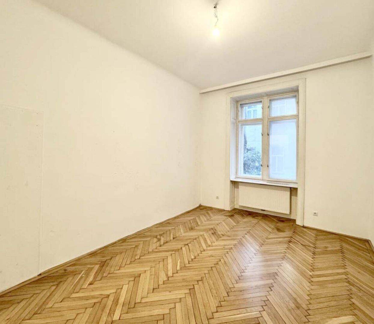 Leerer Raum mit Fischgrätparkettboden und einem Fenster mit Heizkörper darunter.