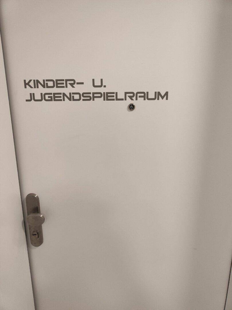 Weiße Tür mit Beschriftung für einen Kinder- und Jugendspielraum in einem Gemeinschaftsbereich.