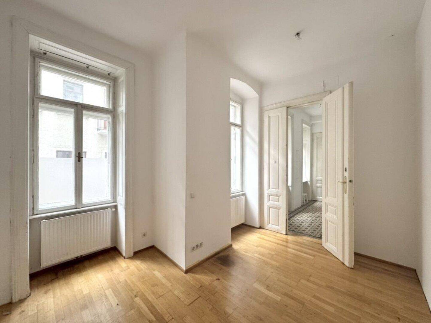 Geräumiges Zimmer mit Parkettboden, hohen Fenstern und Zugang zu einem weiteren Raum.