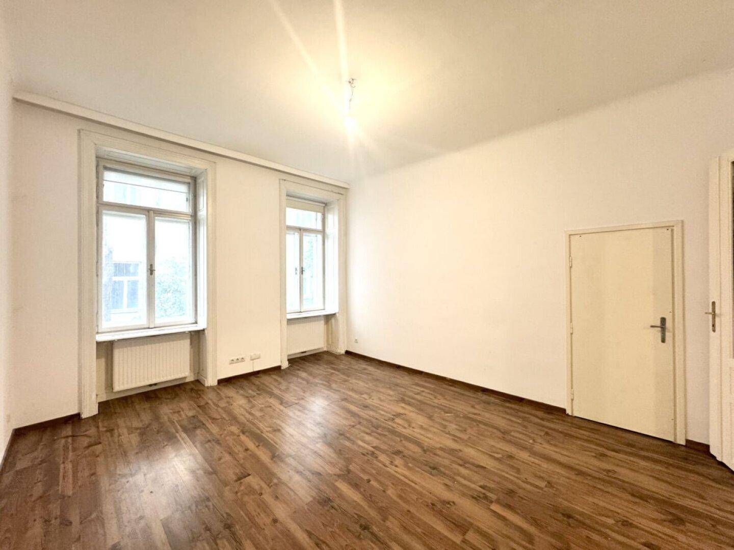 Helles Zimmer mit Holzboden, zwei großen Fenstern und viel Tageslicht.