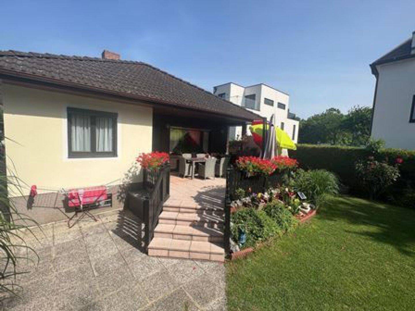Hinterer Bereich des Hauses mit einer gefliesten Terrasse und angrenzendem Garten unter blauem Himmel.