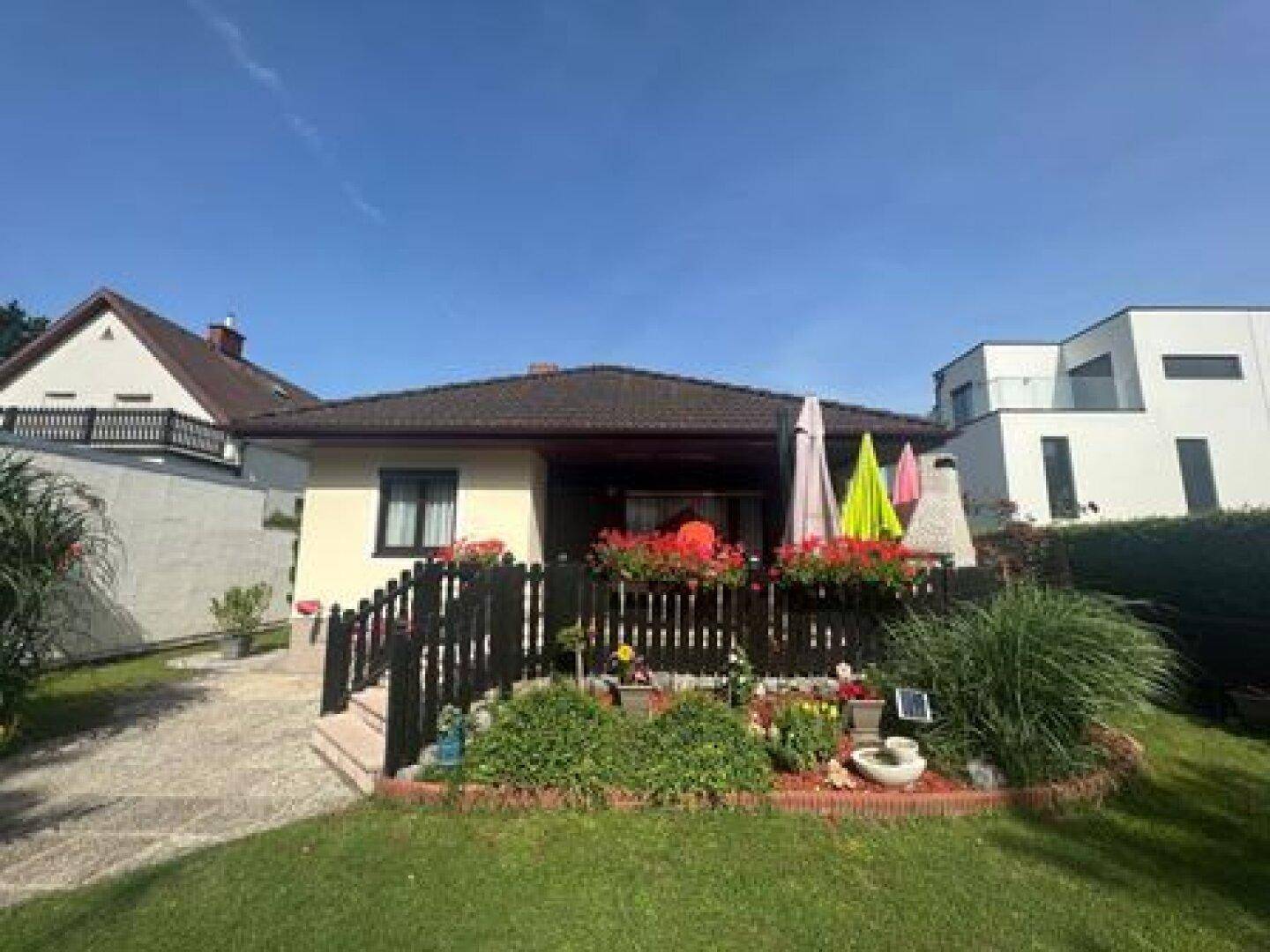 Rückansicht des Hauses mit Terrasse, Blumenbeeten und weitläufiger Rasenfläche unter klarem Himmel.