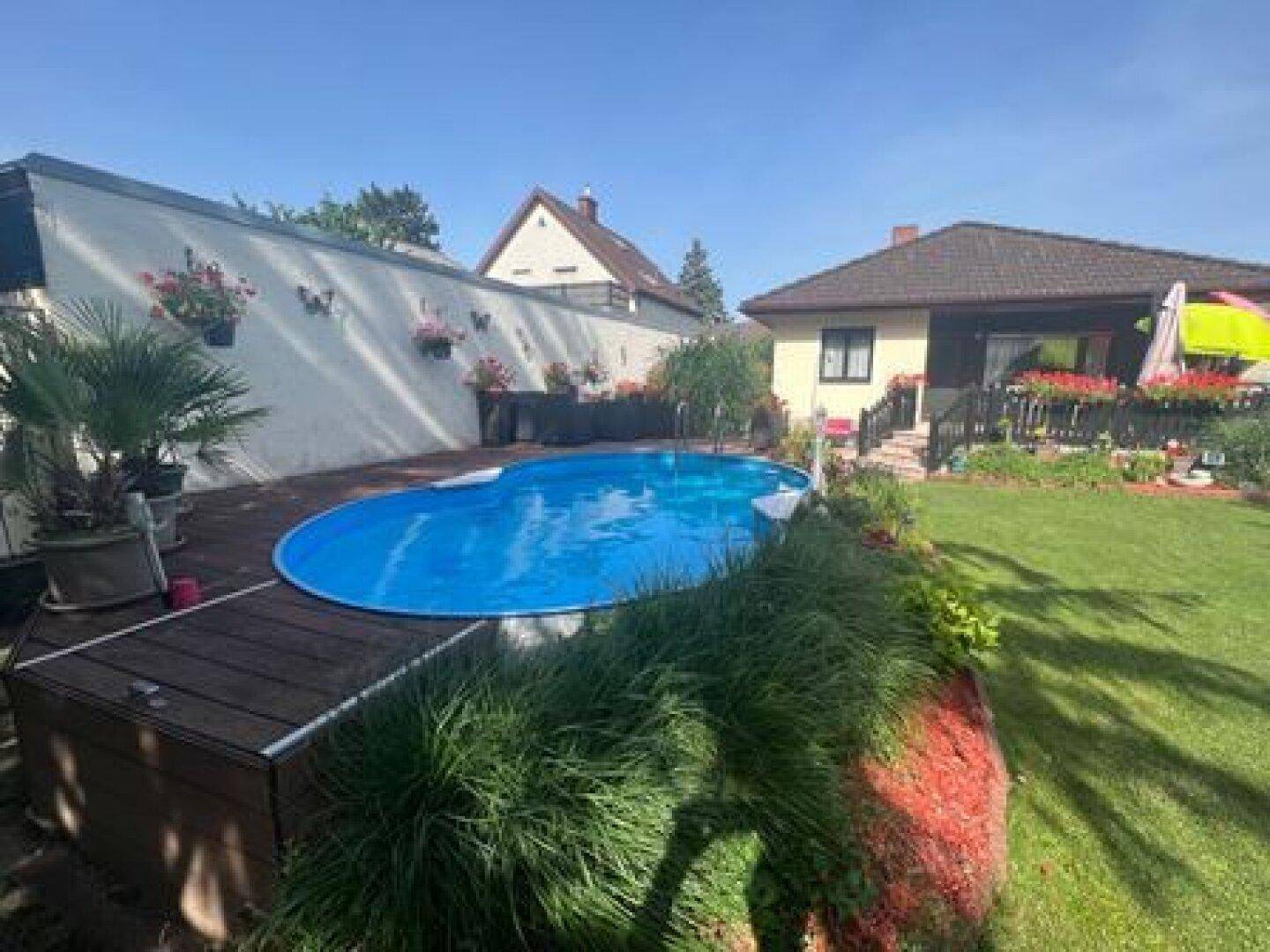 Garten mit ovalem Pool, umgeben von einer Holzterrasse und grünen Pflanzen unter blauem Himmel.