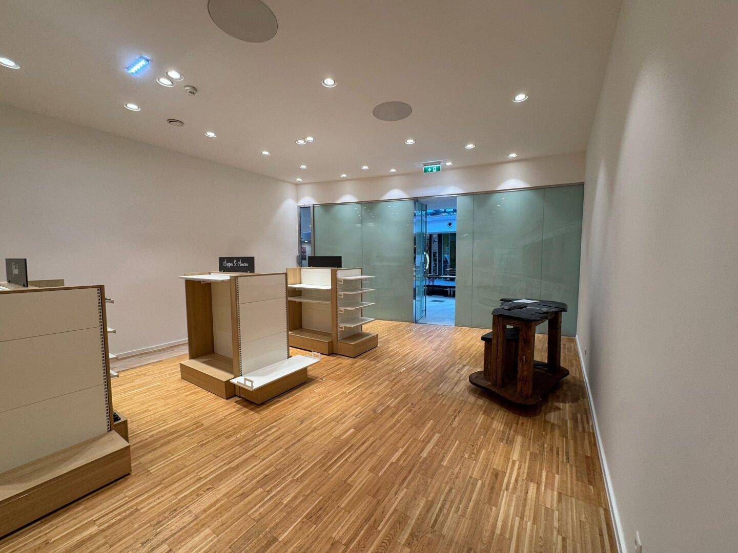 Leeres Ladenlokal mit hellem Holzfußboden, weißen Wänden und modernen Glas-Schiebetüren im Hintergrund.