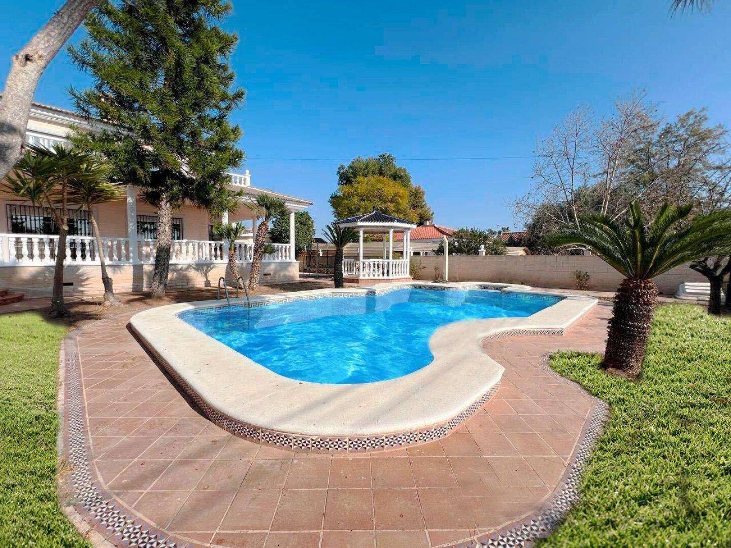 Weitläufige Gartenanlage mit großem Pool, Palmen und mediterraner Bepflanzung unter blauem Himmel.