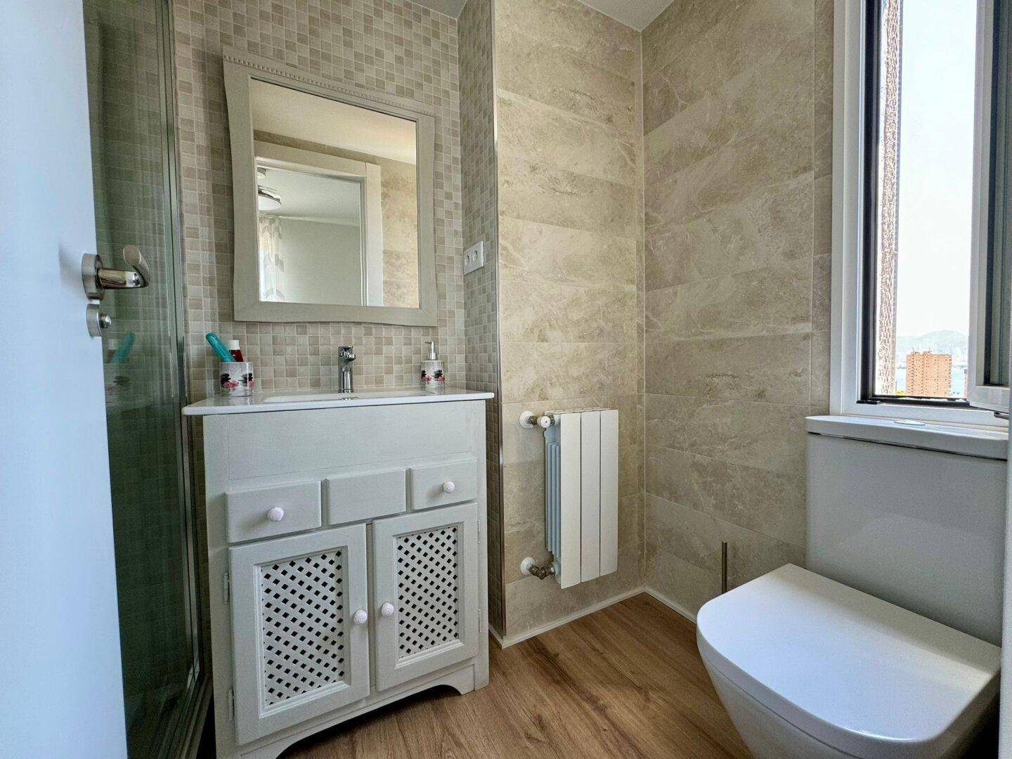 Helles Badezimmer mit Waschtisch, Toilette, Fenster und beigen Wandfliesen.