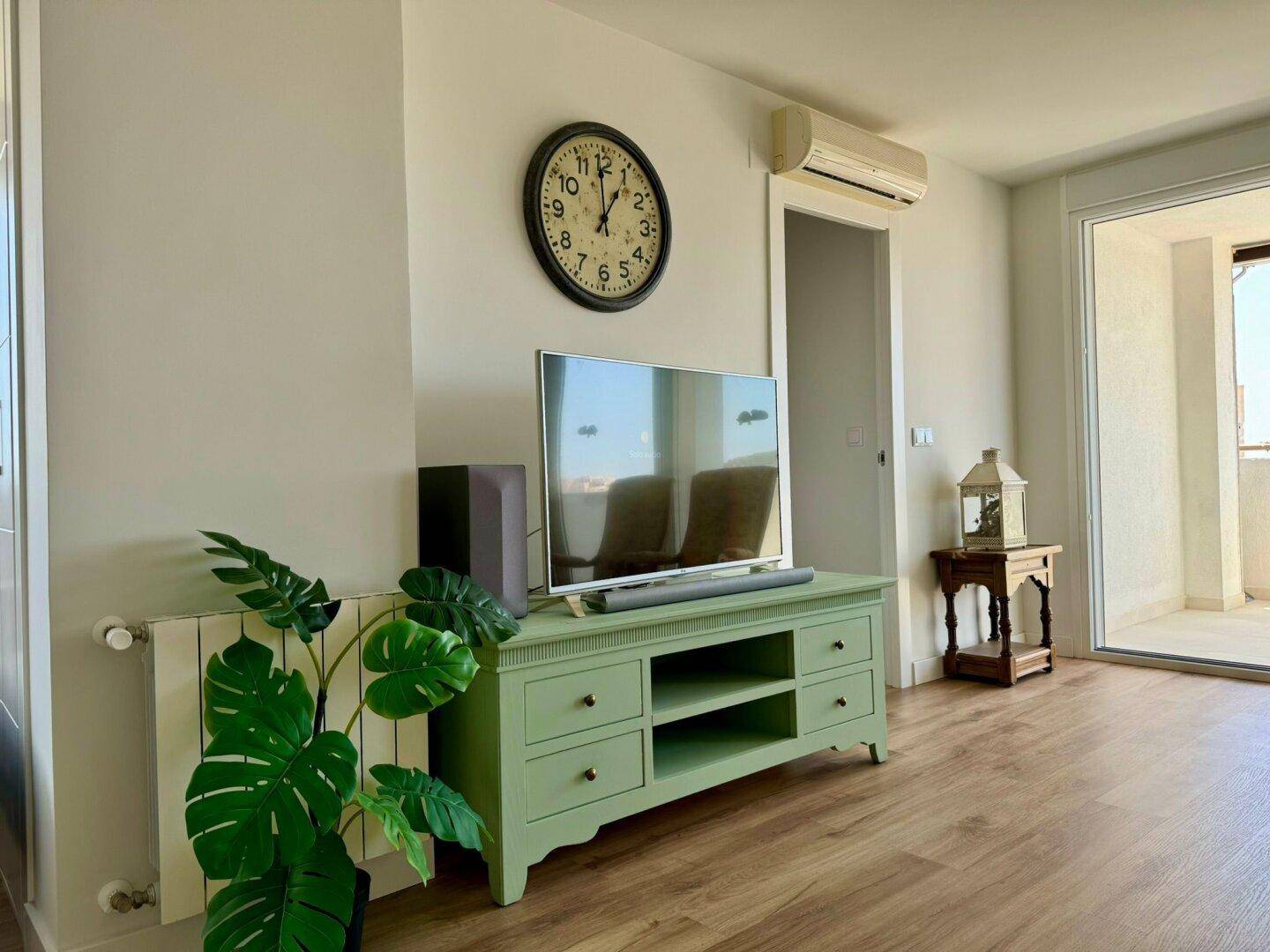 Detailansicht des Wohnbereichs mit TV, grünem Sideboard, Wanduhr und Klimaanlage.