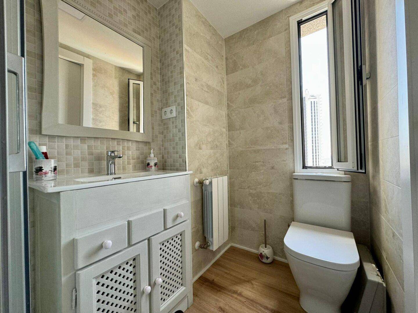 Badezimmer mit Waschtisch, Toilette, Fenster und hellen Fliesen, die für viel Tageslicht sorgen.