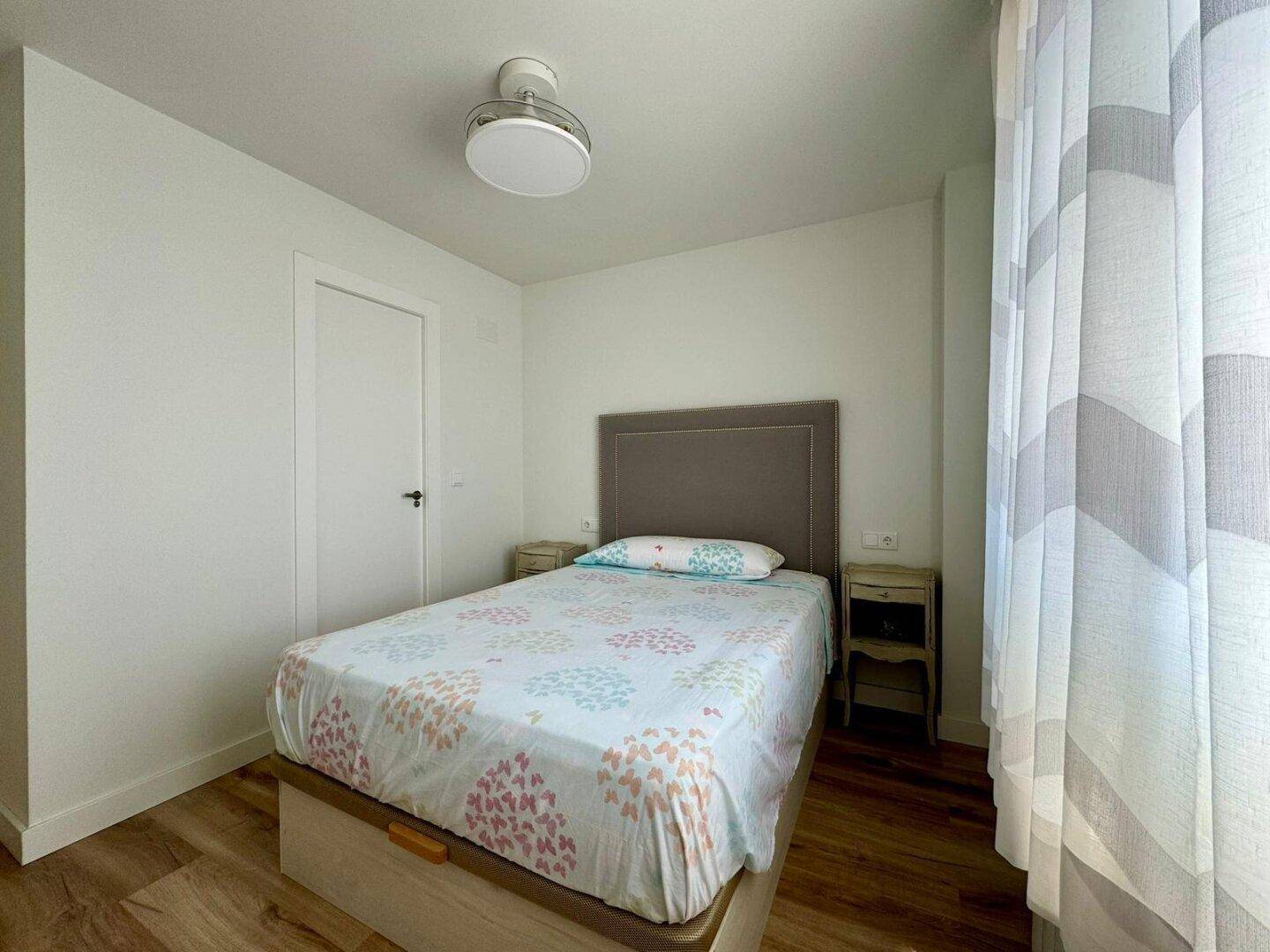 Helles Schlafzimmer mit Einzelbett, Holzboden, Deckenventilator und natürlichem Lichteinfall.