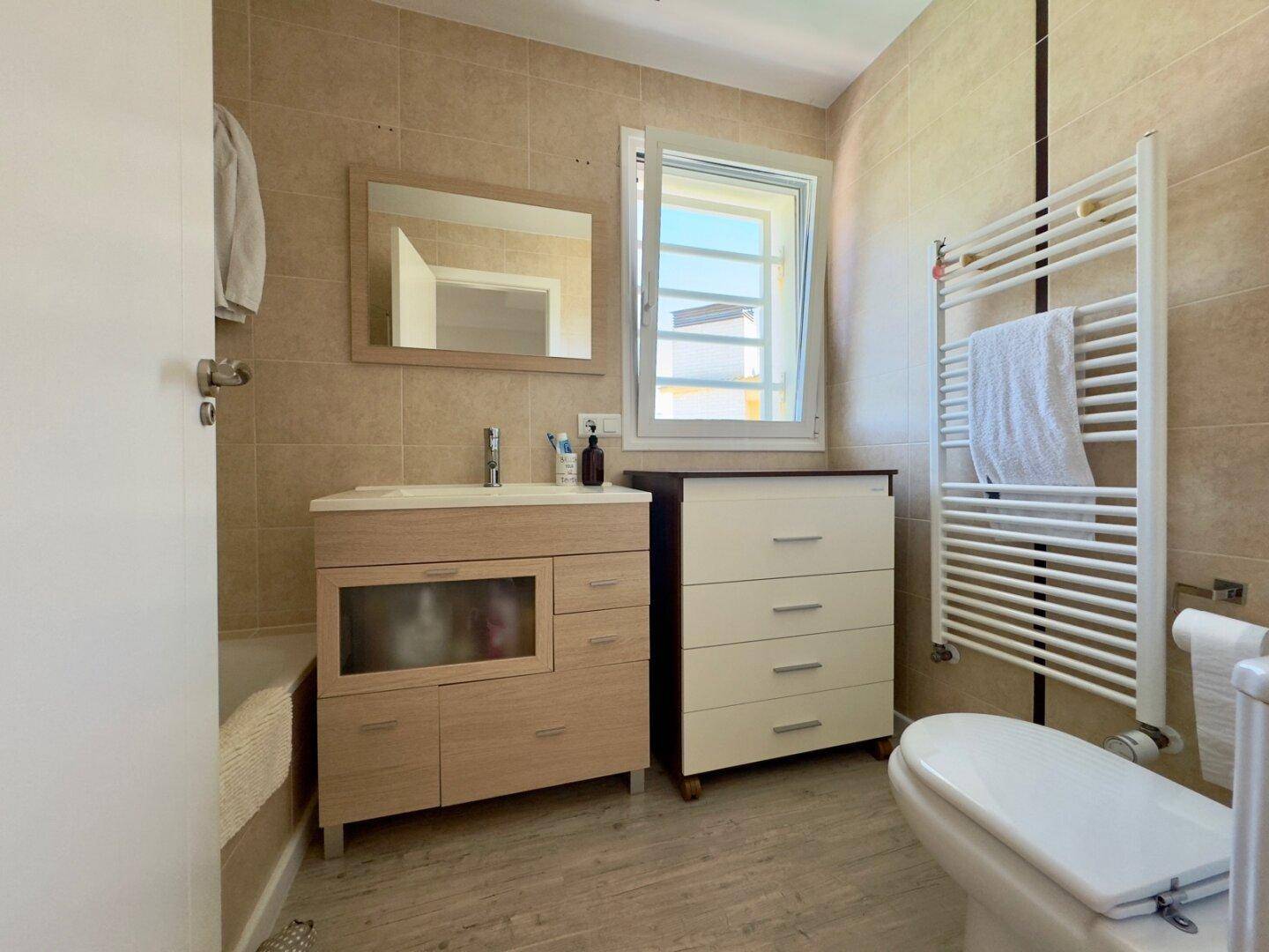 Badezimmer mit Waschtisch, WC, beheiztem Handtuchhalter und Fenster für natürliche Belüftung.