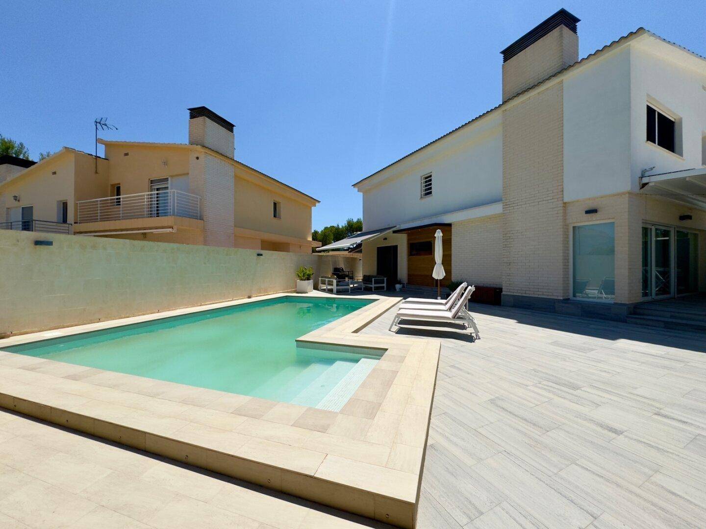 Moderne Villa mit großem Poolbereich und sonniger Terrasse, umgeben von Grün.