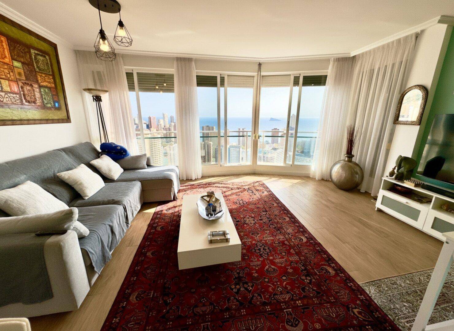 Helles Wohnzimmer mit großen Fenstern und Panoramablick auf Stadt und Meer. Moderner Stil mit Laminatboden und einem farbigen Teppich.