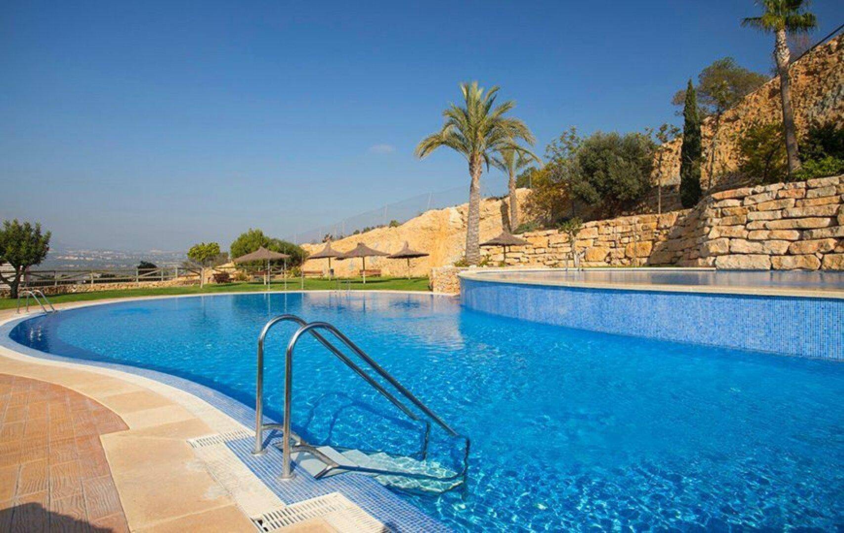 Großes Schwimmbecken mit blauen Mosaikfliesen, umgeben von Palmen und einer natürlichen Felswand.
