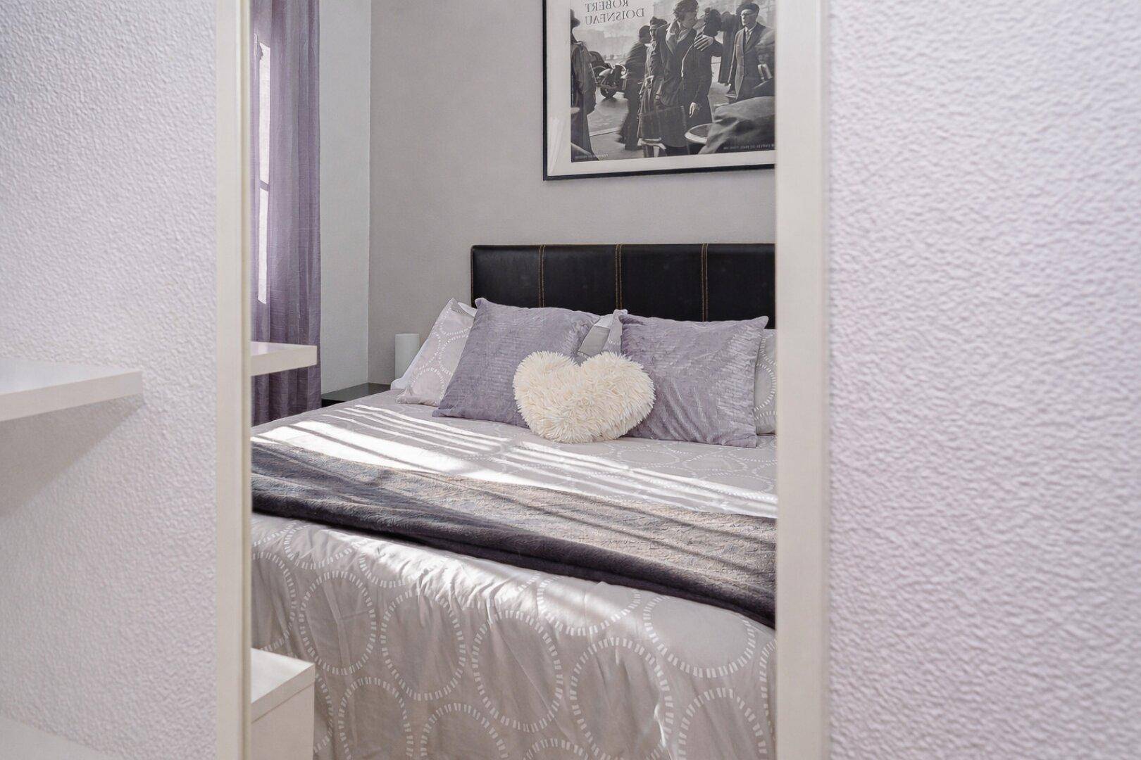Gemütliches Schlafzimmer mit Doppelbett und lila Vorhängen, durch das Fenster fällt viel Licht.
