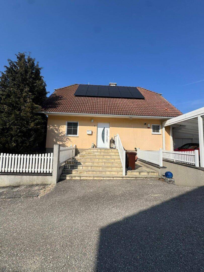 Frontansicht des Einfamilienhauses mit weißem Zaun, Treppe zum Eingang und Carport.