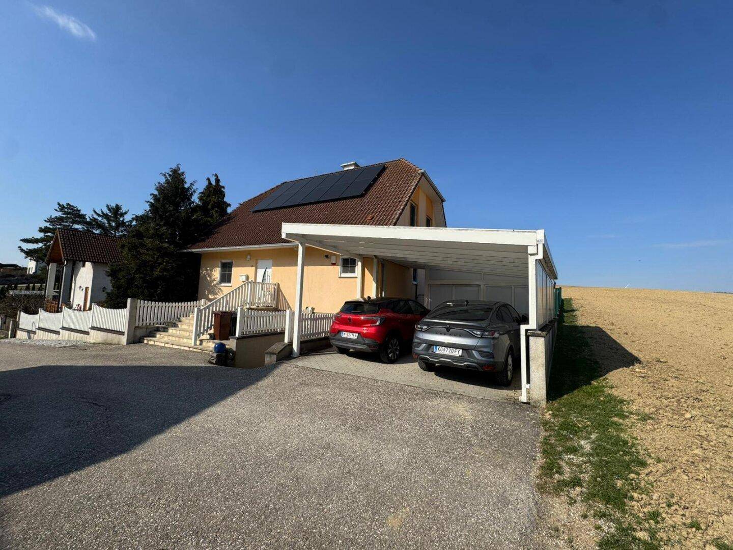 Das Haus mit Carport und zwei geparkten Autos, angrenzend an ein offenes Feld.