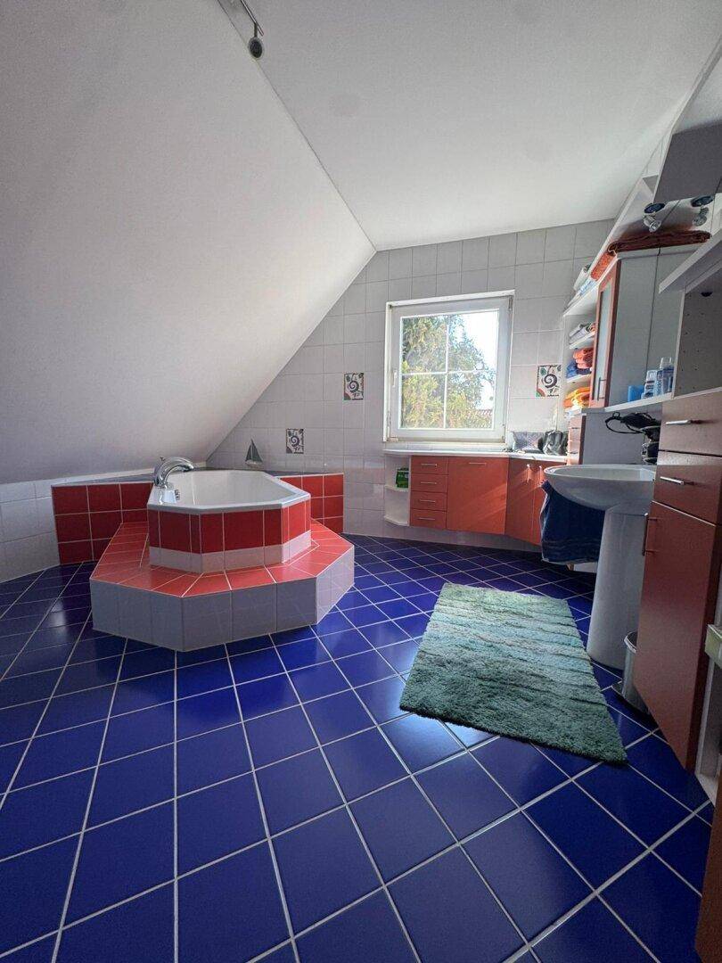 Dachgeschoss-Badezimmer mit Eckbadewanne, blauen Fliesen und einem Fenster, das viel Licht hereinlässt.