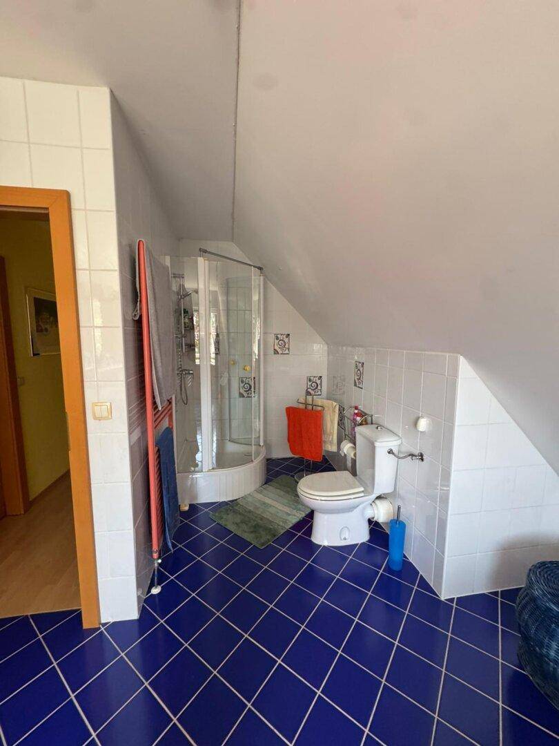 Dachgeschoss-Badezimmer mit Duschkabine, WC und blauen Fliesen, unter einer Dachschräge gelegen.