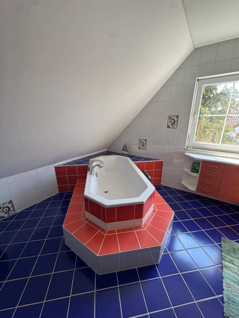 Dachgeschoss-Badezimmer mit Eckbadewanne, blauen Fliesen und einem Fenster, das den Raum mit Tageslicht versorgt.