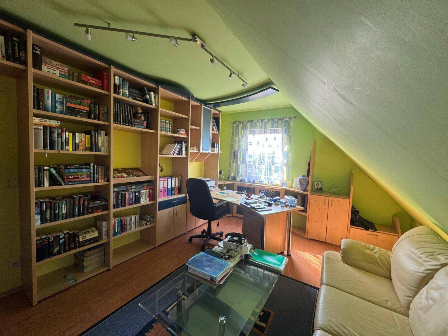 Dachgeschoss-Büro mit eingebauten Bücherregalen, Schreibtisch und Sofa, unter einer grünen Dachschräge.