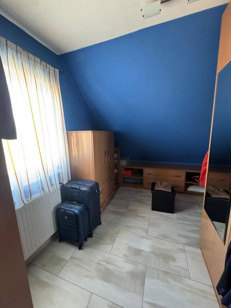 Dachgeschoss-Schlafzimmer mit blauer Akzentwand, Kleiderschrank und Fenster, unter einer Dachschräge.