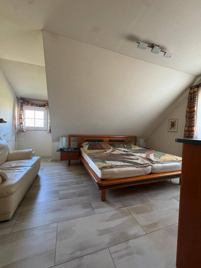 Dachgeschoss-Schlafzimmer mit Doppelbett, Sofa und Fenster, das den Raum mit Licht füllt.