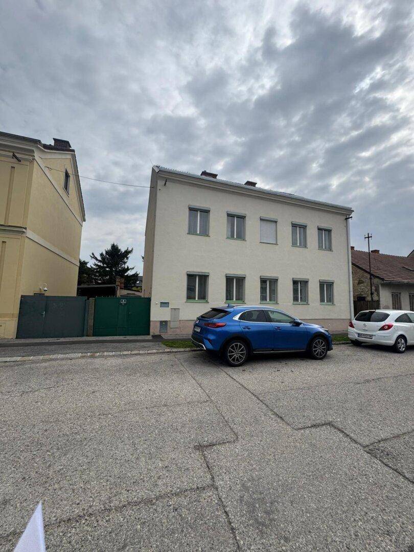 Ansicht der Gebäudefassade von der Straße mit parkenden Autos und bewölktem Himmel.