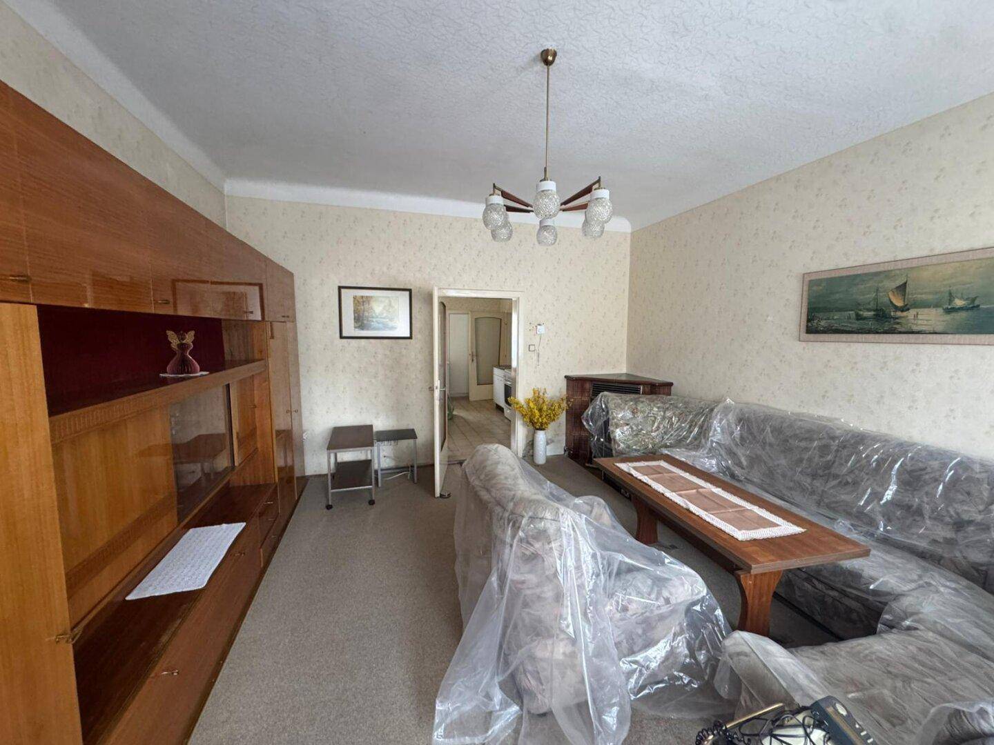 Wohnzimmer mit Teppichboden, Holzmöbeln und abgedeckten Sofas, bereit zur Renovierung.