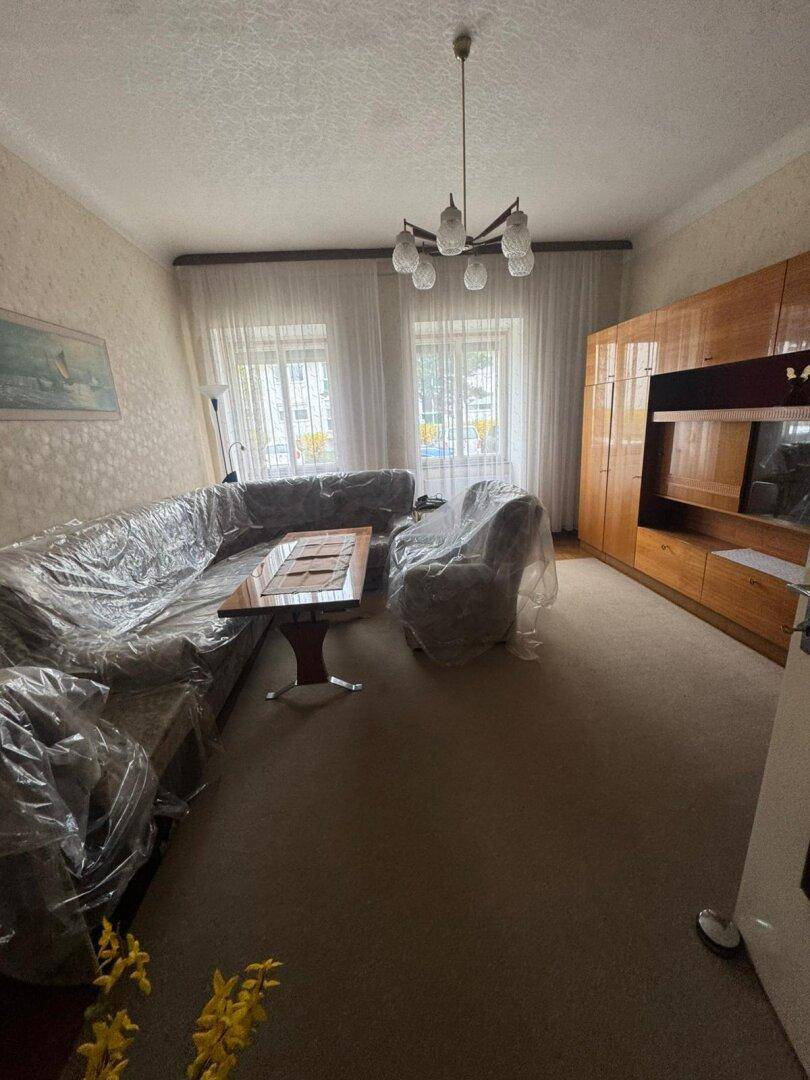 Wohnzimmer mit Teppichboden, großen Fenstern und abgedeckten Möbeln, die auf eine Renovierung warten.