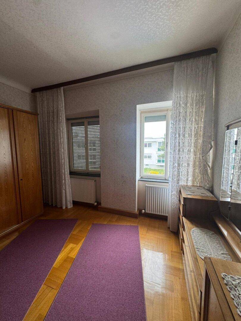 Schlafzimmer mit zwei Fenstern, Parkettboden und Holzmöbeln.