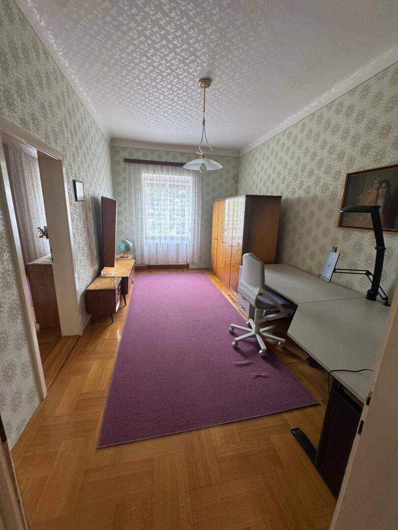 Schlafzimmer mit Schreibtisch, Parkettboden und gemusterter Tapete.