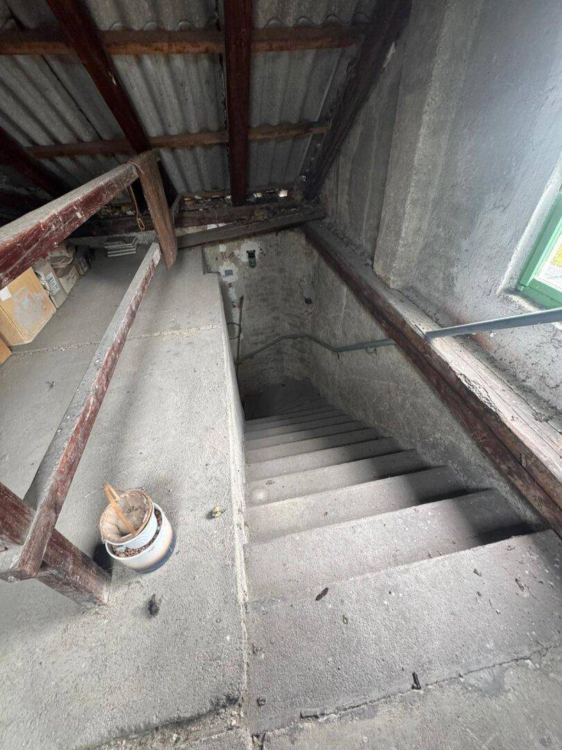 Steile Betontreppe führt in die Tiefe, umgeben von rohen Wänden und sichtbaren Dachbalken.