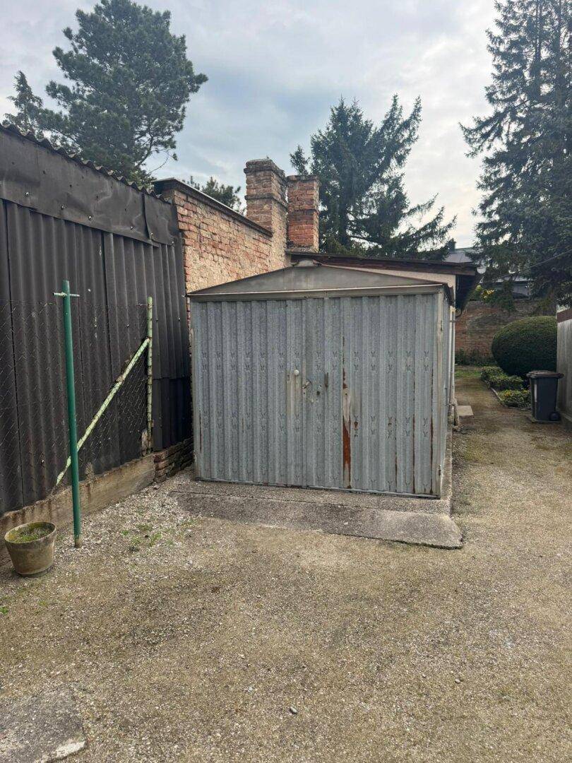 Wellblechschuppen im Gartenbereich, umgeben von Bäumen und einer Ziegelmauer.