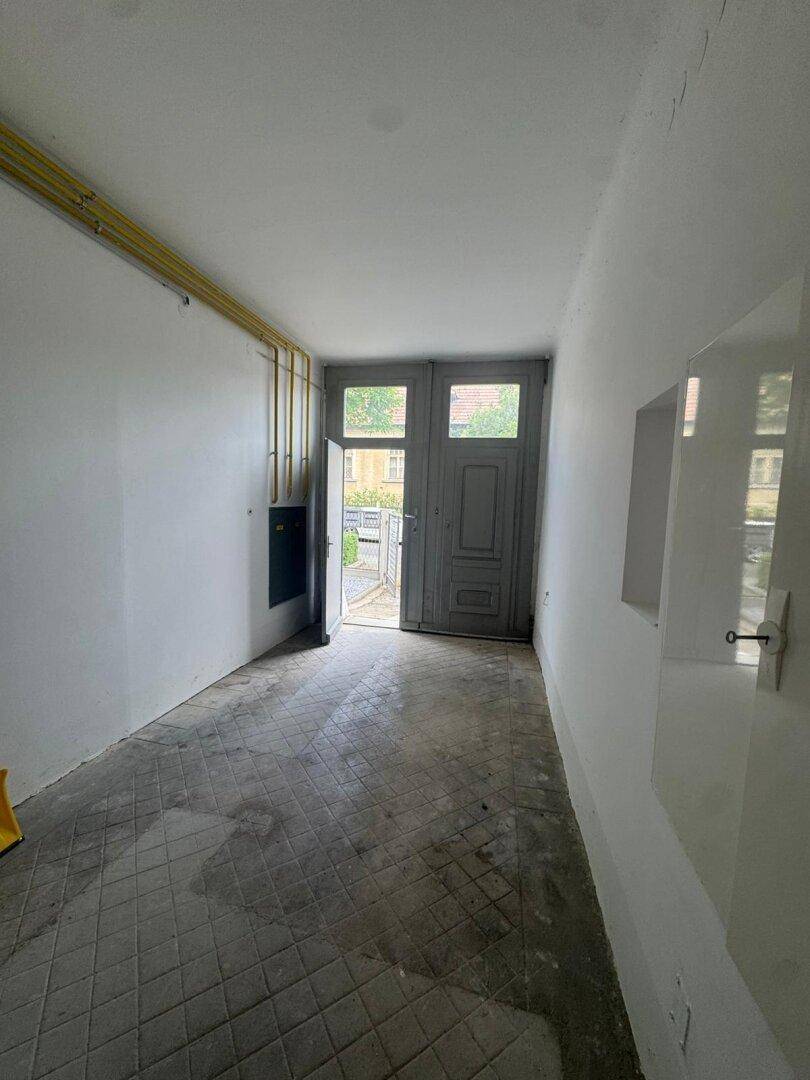 Schmaler Flur oder Abstellraum mit gefliestem Boden und einer alten Holztür, die nach draußen führt.