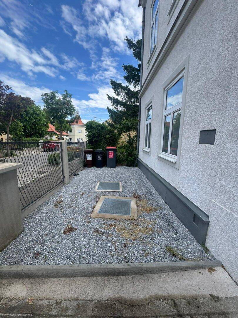 Außenbereich vor dem Gebäude mit Kiesfläche, Mülltonnen und Zugangspunkten am Fundament.
