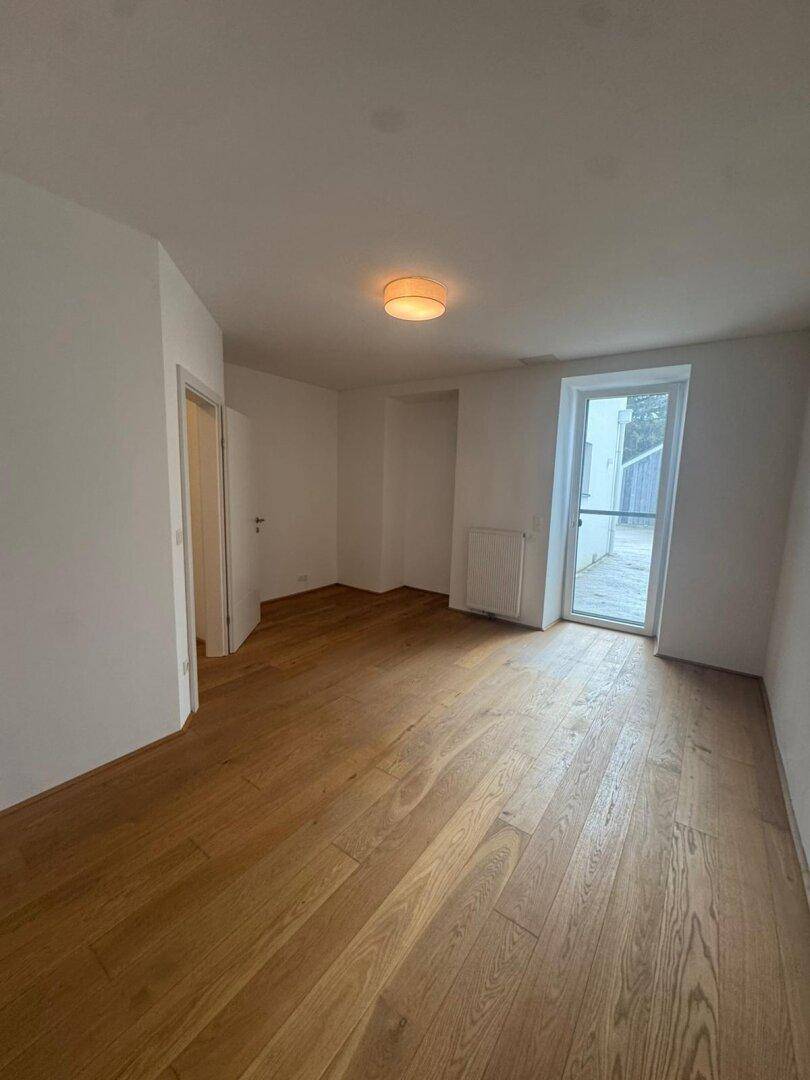 Geräumiger Raum mit Hartholzboden, weißen Wänden und Zugang zu einem Außenbereich.