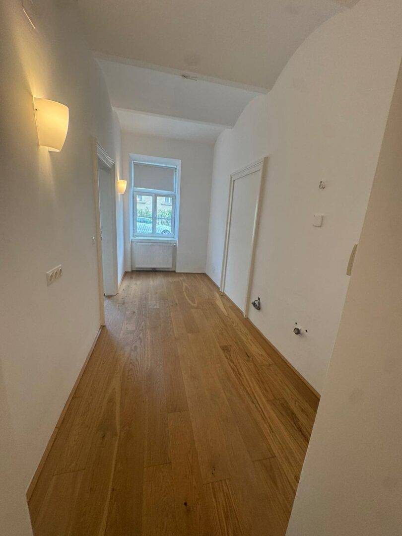 Heller Raum mit neuem Hartholzboden, weißen Wänden und einem Fenster, das Tageslicht hereinlässt.