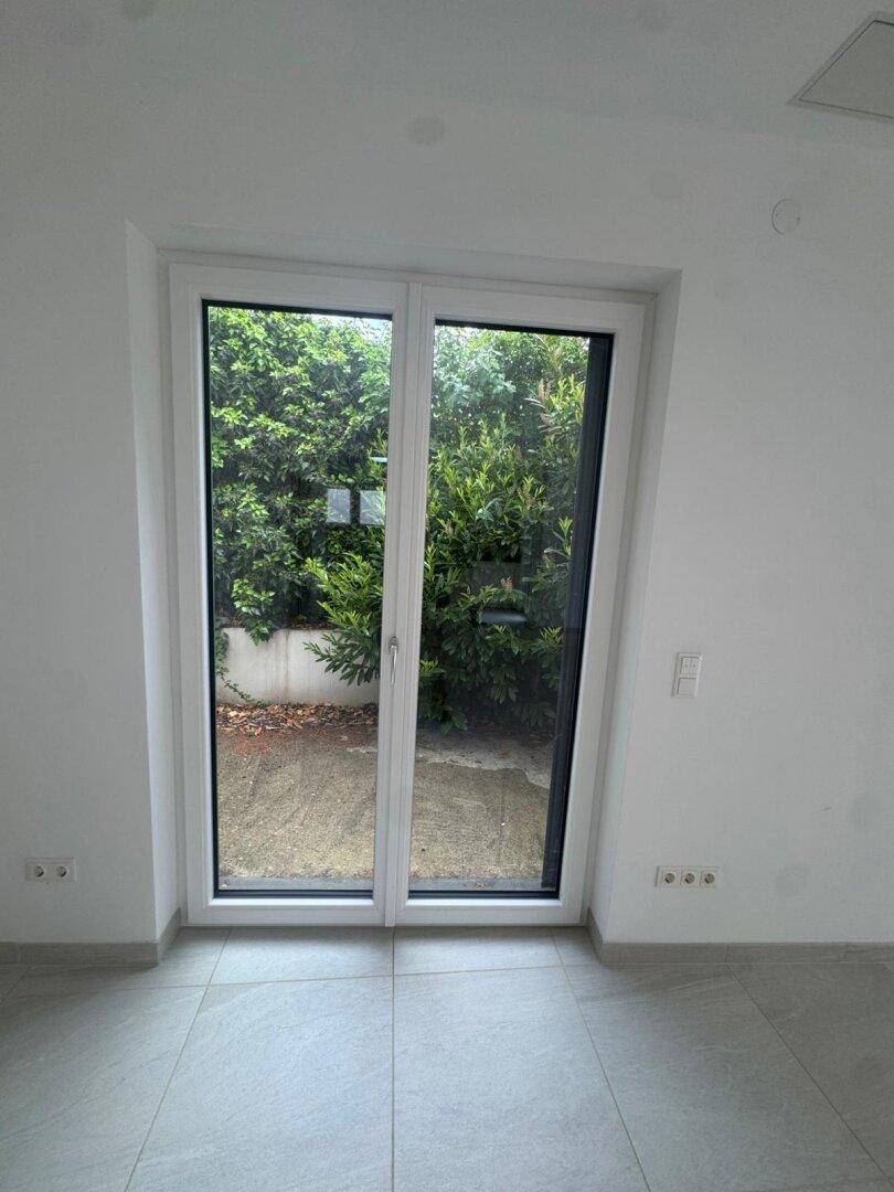 Große Glastür mit Zugang zum Garten, umgeben von grünen Büschen und hellem Fliesenboden.