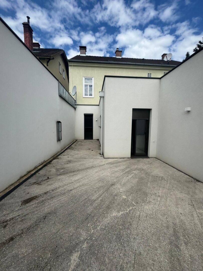 Schmaler Innenhof mit Betonboden zwischen weißen Gebäuden, der zu einem weiteren Eingang führt.