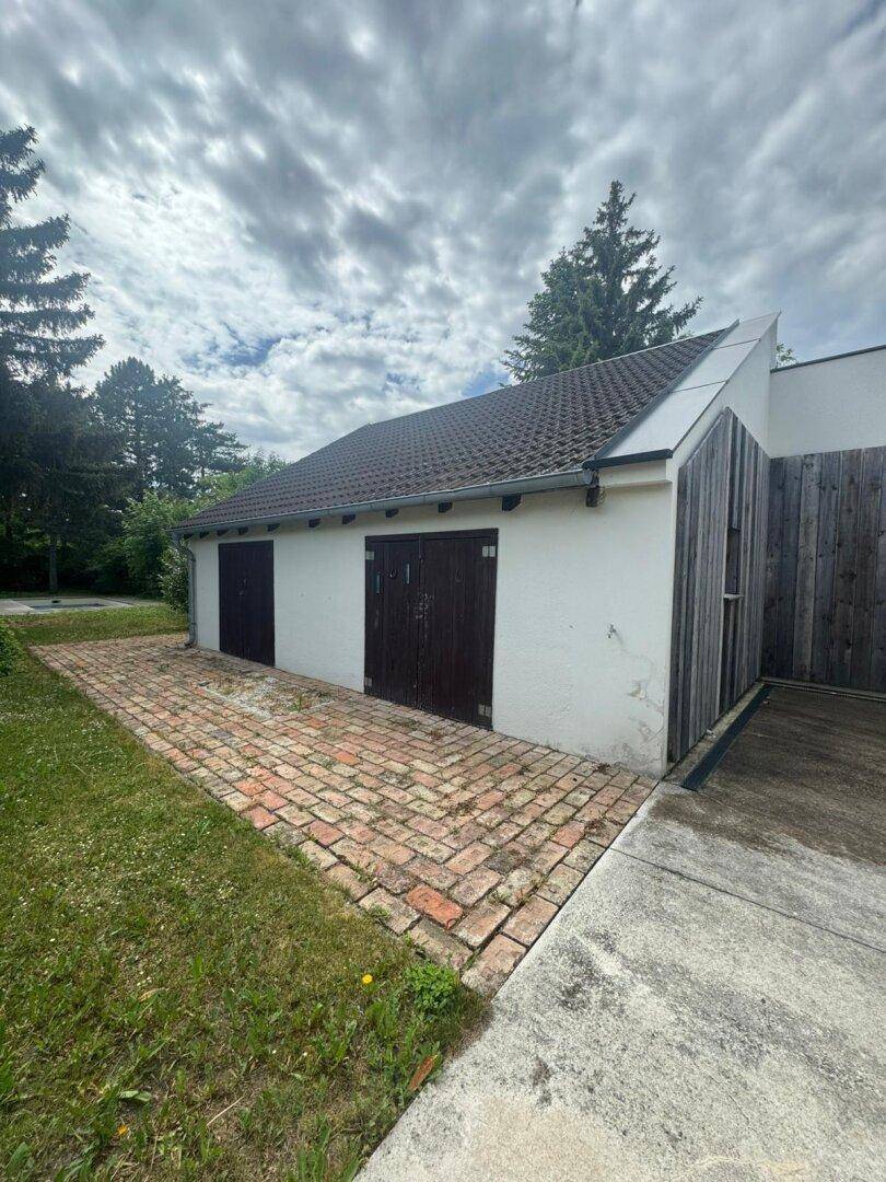 Älteres Nebengebäude mit zwei dunklen Garagentoren, umgeben von Rasen und Bäumen.
