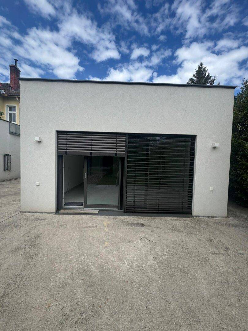 Moderne weiße Fassade des Anbaus mit großen Fenstern und Jalousien unter klarem Himmel.