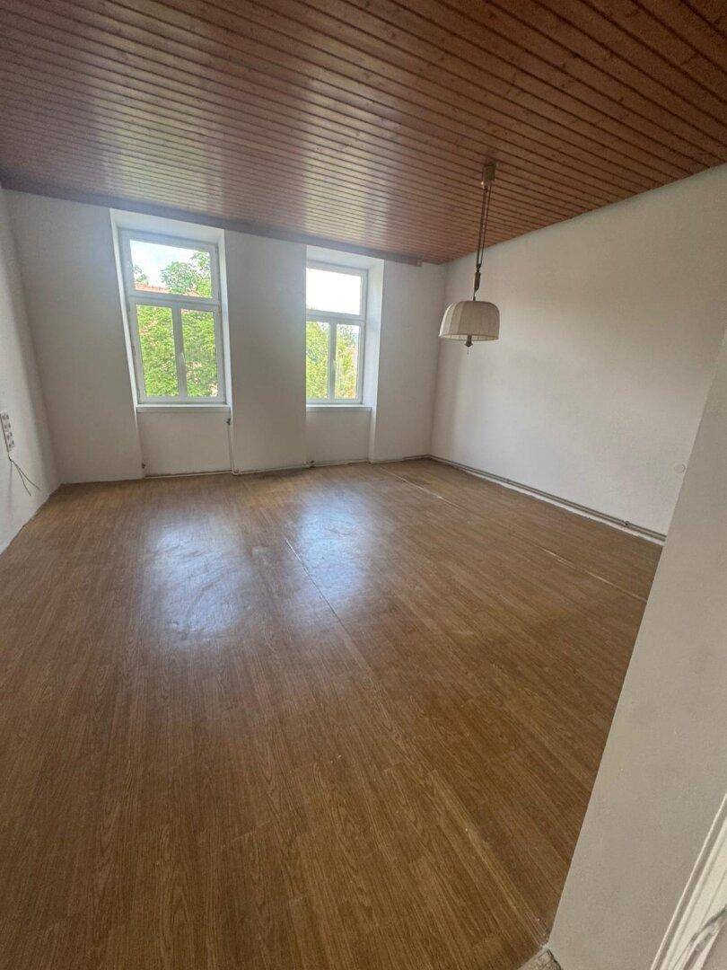 Geräumiger Raum mit Holzboden, Holzpaneeldecke und zwei Fenstern, die Tageslicht hereinlassen.