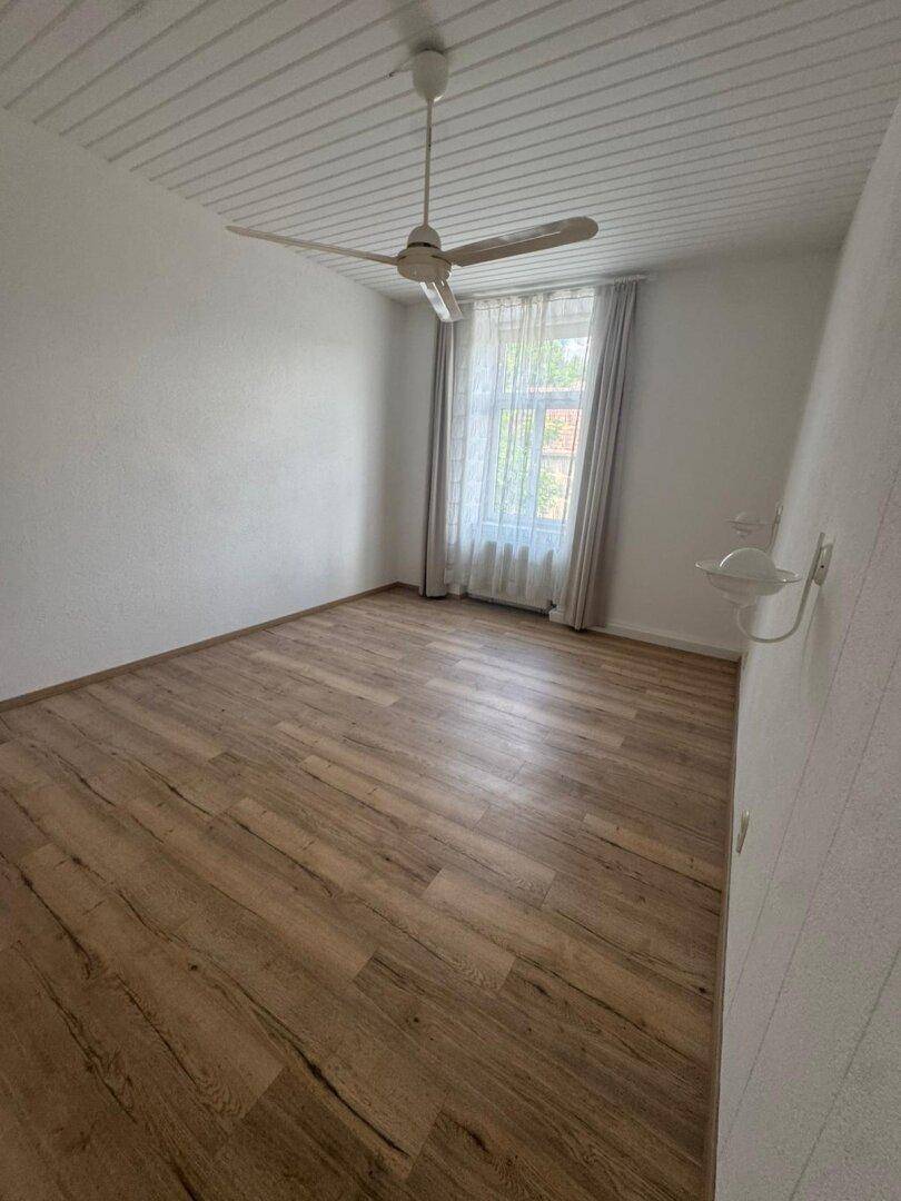 Schlafzimmer mit Holzboden, weißer Paneeldecke, Fenster mit Vorhängen und Deckenventilator.