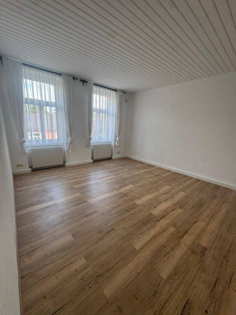 Helles Schlafzimmer mit Holzboden, weißer Paneeldecke und zwei Fenstern mit Heizkörpern.