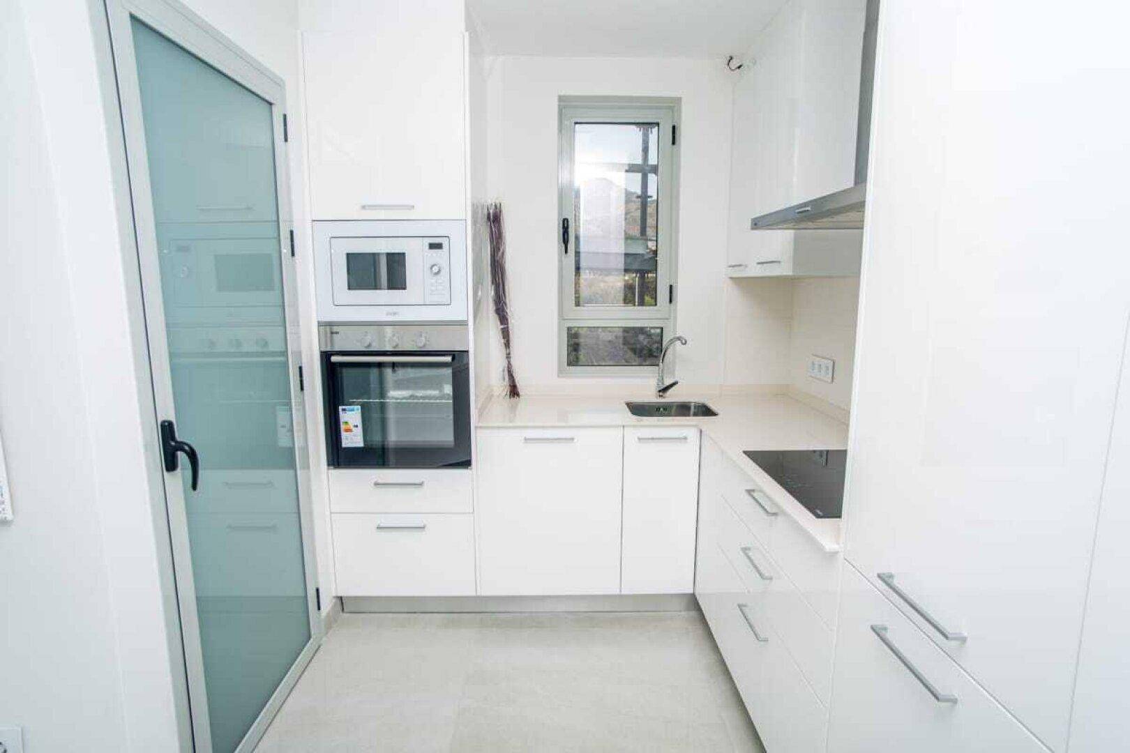 Moderne weiße Küche mit Einbaugeräten, Fenster und heller Arbeitsplatte.