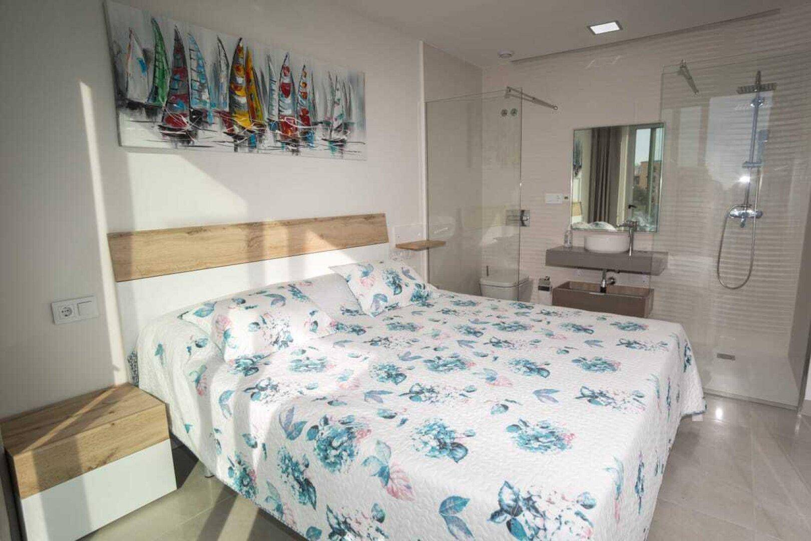 Helles Schlafzimmer mit floraler Bettwäsche und offenem Badbereich mit Dusche.